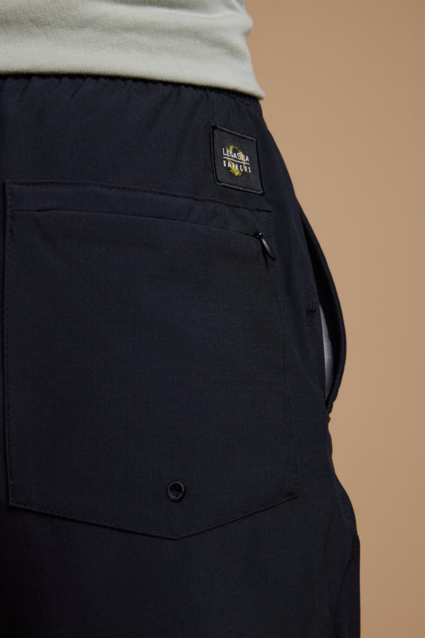 Close-up of the Legasea Weekend Short by Legasea, showing the recycled polyester back pocket with a button detail and a rectangular patch label, worn with a light shirt against a plain beige background.