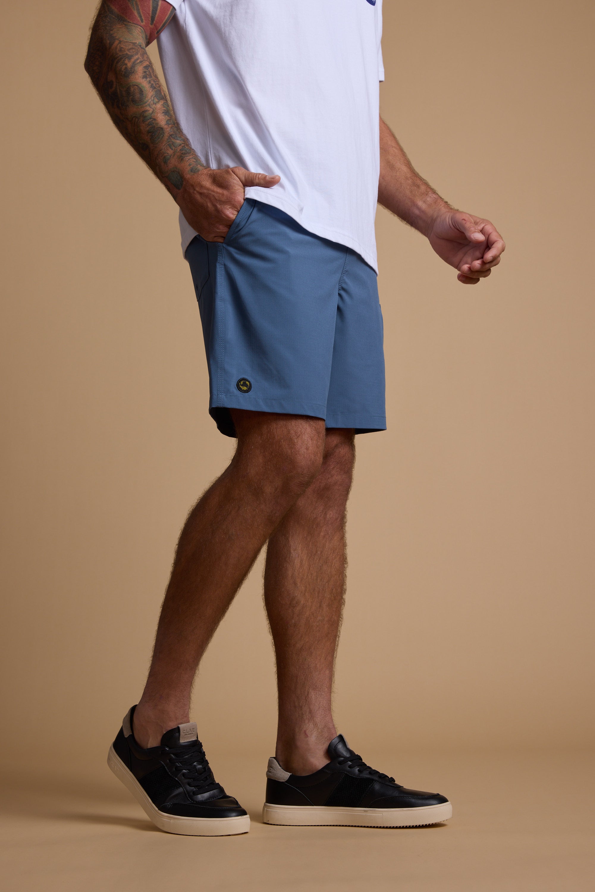 A man wearing a white T-shirt, Legasea Weekend Short in blue, and black sneakers with white soles stands against a beige background. His left hand is in his pocket, tattoos visible on his left arm. His face is not shown.