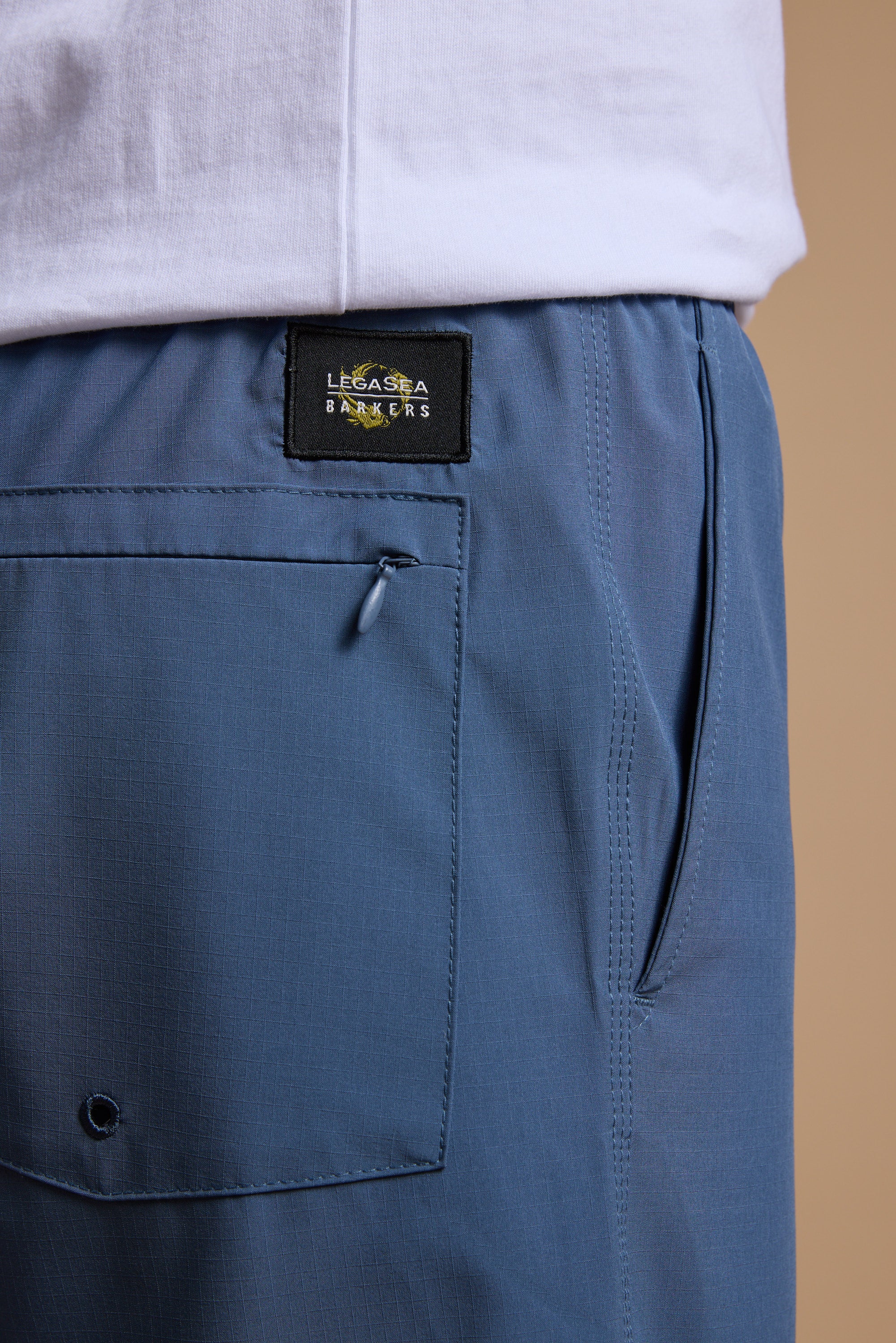 Close-up of a person wearing Legasea Weekend Shorts in blue recycled polyester with a back pocket and black Legasea label, paired with a white shirt, set against a neutral beige background.