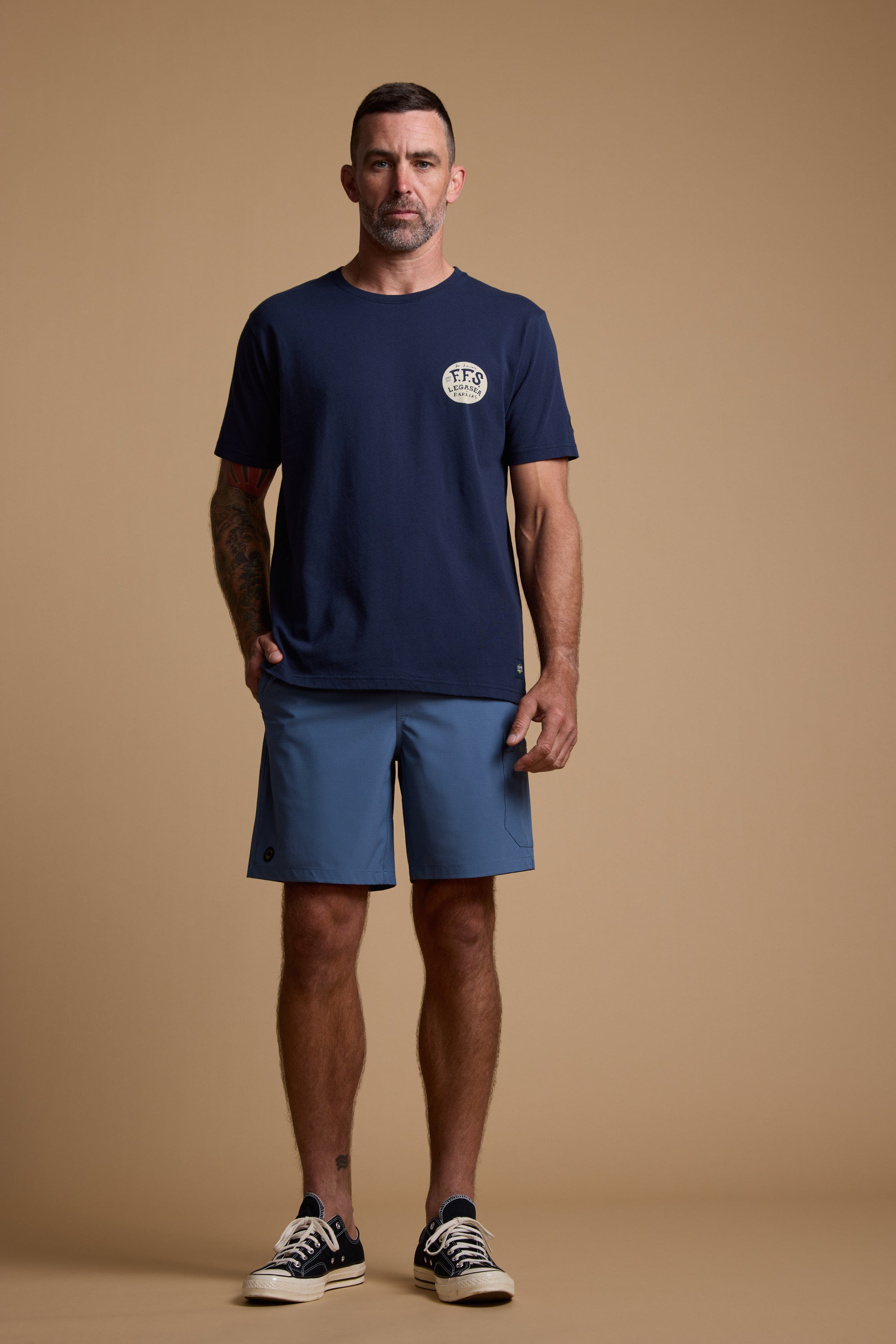 A man stands against a beige background wearing a navy T-shirt, Legasea Weekend Shorts in blue, and black-and-white sneakers. His right hand is in his pocket and he has a neutral expression.