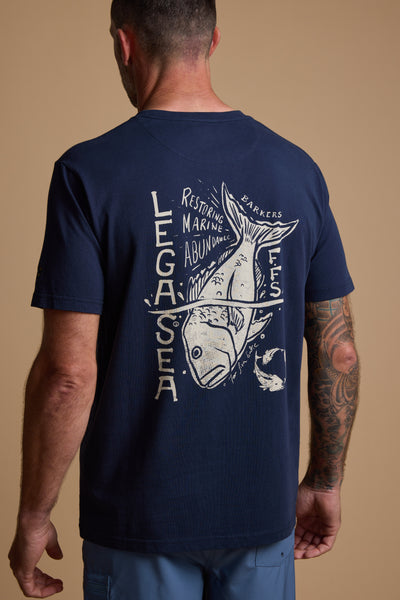 A man with a tattooed right arm wears a navy Legasea Snapper Print Tee by Legasea, featuring a large fish graphic and text inspired by the New Zealand coastline.