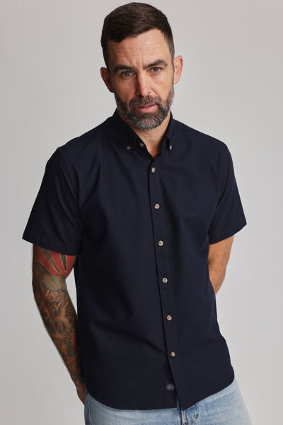A man with short dark hair, a beard, and a tattooed right arm wears the Barkers Archer Oxford Shirt in navy with a button-down collar and light blue jeans, standing against a plain light background.
