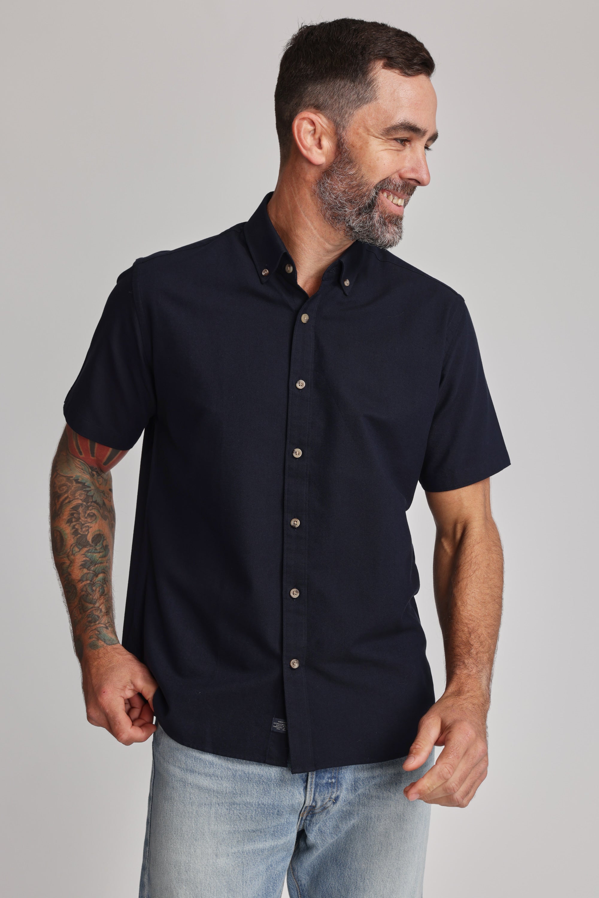 A man with short dark hair, a beard, and a tattooed arm wears the Barkers Archer Oxford Shirt in navy with a button-down collar and light blue jeans, smiling and looking to the side against a plain background.