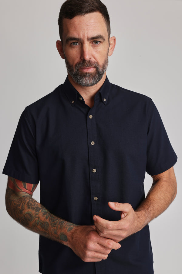 A man with short dark hair, a beard, and visible tattoos on his left arm wears the Barkers Archer Oxford Shirt, a 100% cotton navy button-down. He stands against a plain light background, looking directly at the camera.