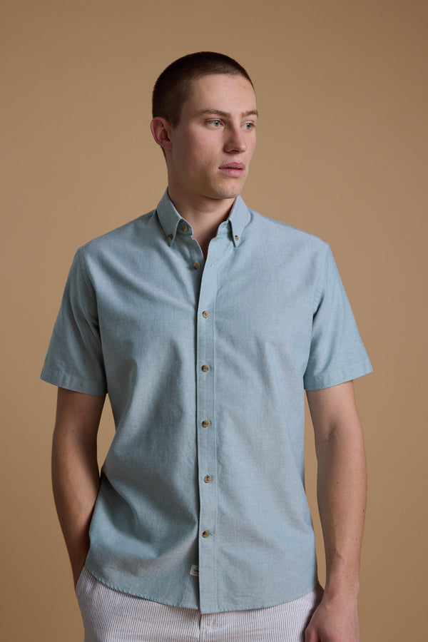 A young man with short hair wears the Barkers Archer Oxford Shirt, made from 100% cotton in light blue with a button-down collar, paired with light striped pants, standing against a beige background and looking to his left.