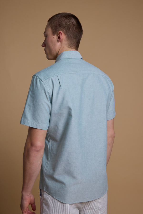 A man with short hair is seen from behind wearing the Barkers Archer Oxford Shirt in light blue, featuring 100% cotton and a button-down collar, paired with light striped shorts, standing against a plain tan background.