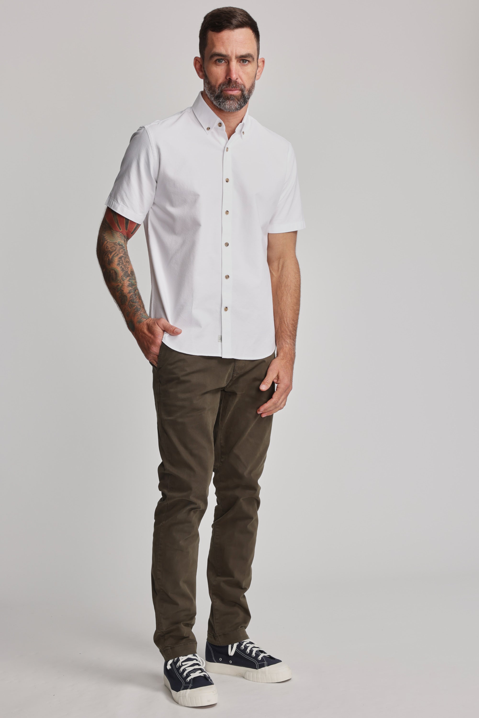 A man with short dark hair and a beard, showing tattoos on his left arm, stands against a plain background wearing the Barkers Archer Oxford Shirt in white with a button-down collar, olive green pants, and black sneakers.