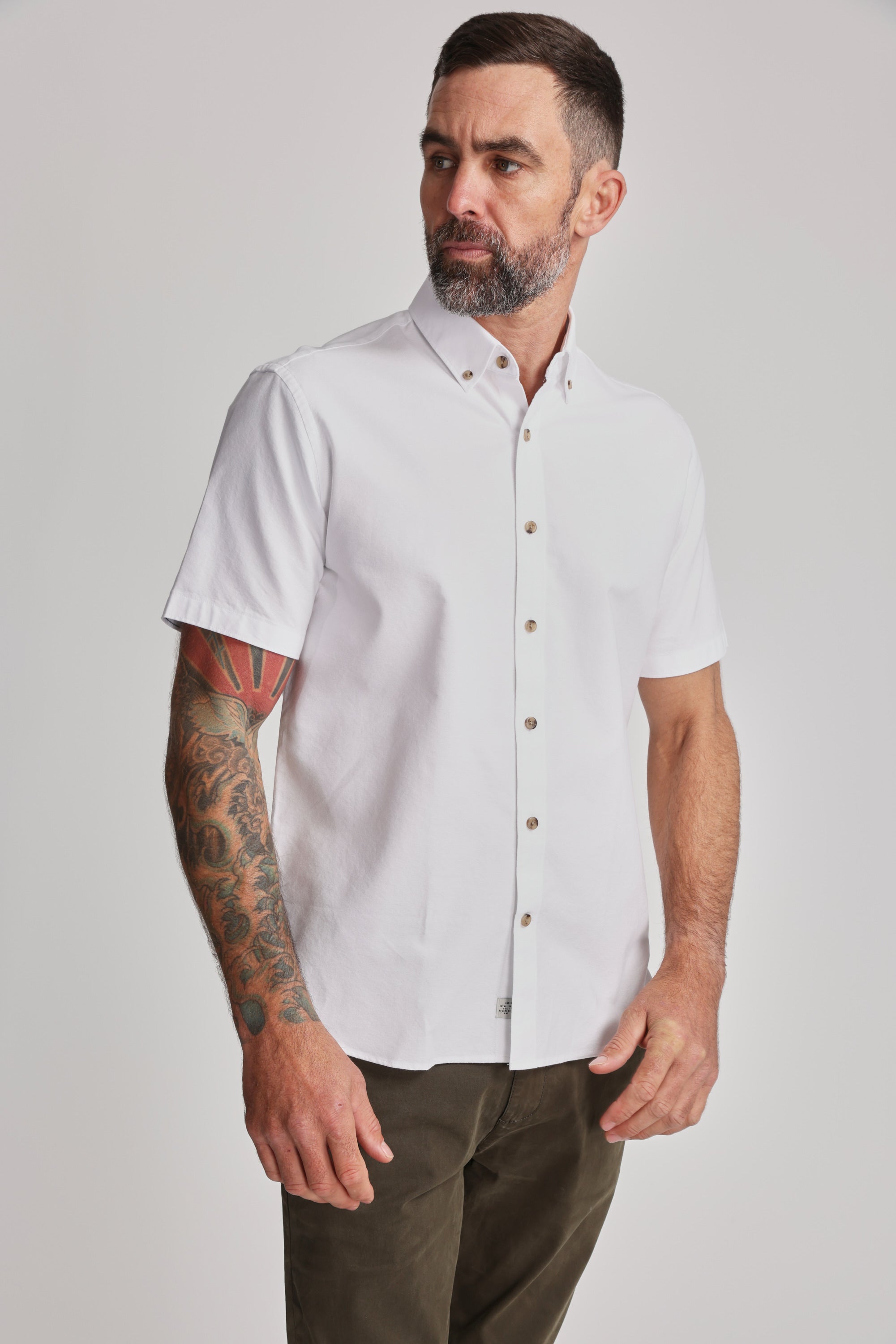 A bearded man with short hair models the Barkers Archer Oxford Shirt—100% cotton, white, with a button-down collar—paired with dark pants. He sports a colorful right-arm tattoo sleeve and gazes slightly left against a plain background.