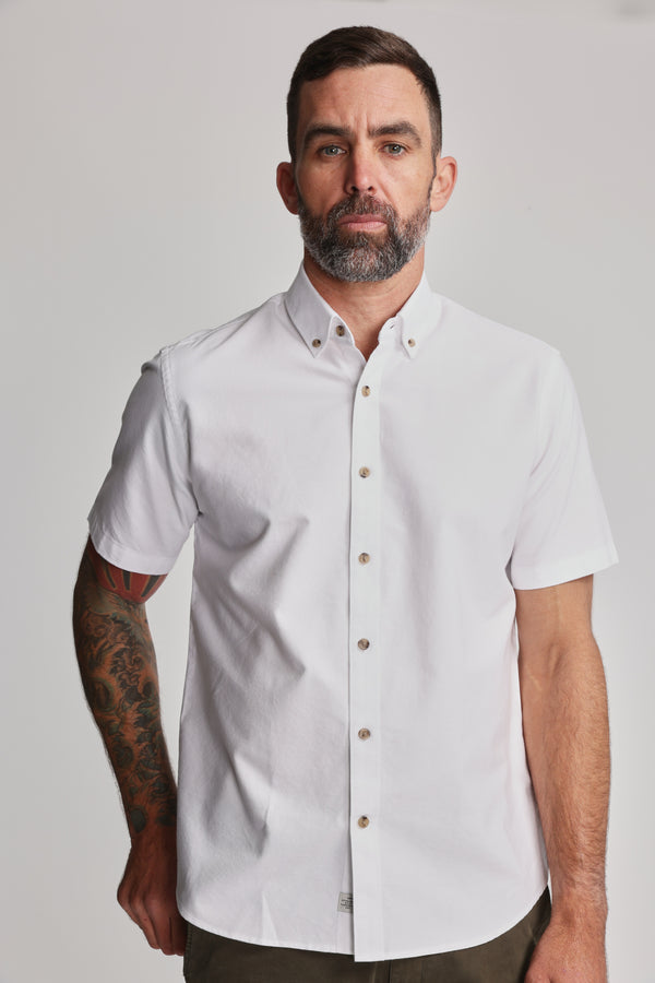 A man with short dark hair and a beard stands facing the camera in a white Barkers Archer Oxford Shirt with a button-down collar. He has a tattoo sleeve on his left arm, a neutral expression, and a plain, light-colored background.