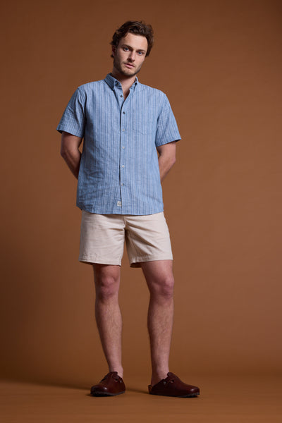 A man stands against a brown backdrop, wearing the Barkers Garvey Stripe Shirt with beige shorts and brown slip-on shoes. His hands are behind his back, and he has a neutral expression.