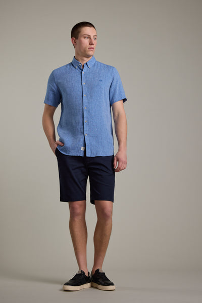 A man stands against a plain background wearing the Barkers Gentry Linen Shirt in light blue, dark navy shorts, and black sneakers with white soles. He has one hand in his pocket and looks to the side.