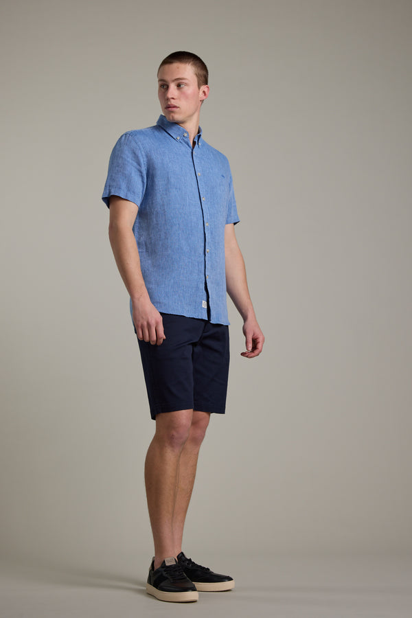 A man stands against a plain background wearing the Barkers Gentry Linen Shirt in light blue, paired with dark navy shorts and black sneakers with white soles, looking slightly to the side.