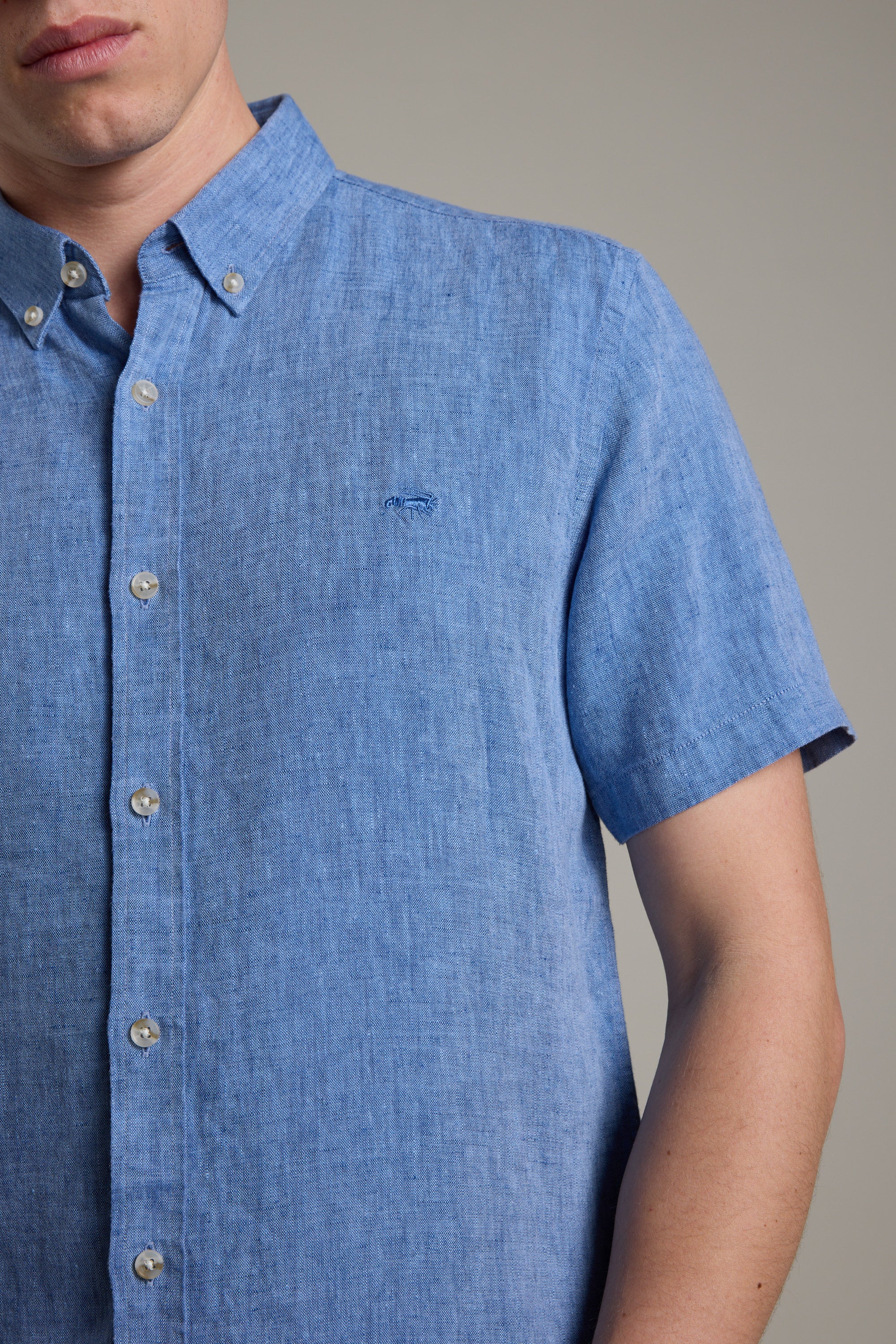 A model wears the Barkers Gentry Linen Shirt in light blue, featuring short sleeves and a small crayfish embroidery on the chest, shown from shoulders to waist against a plain background.