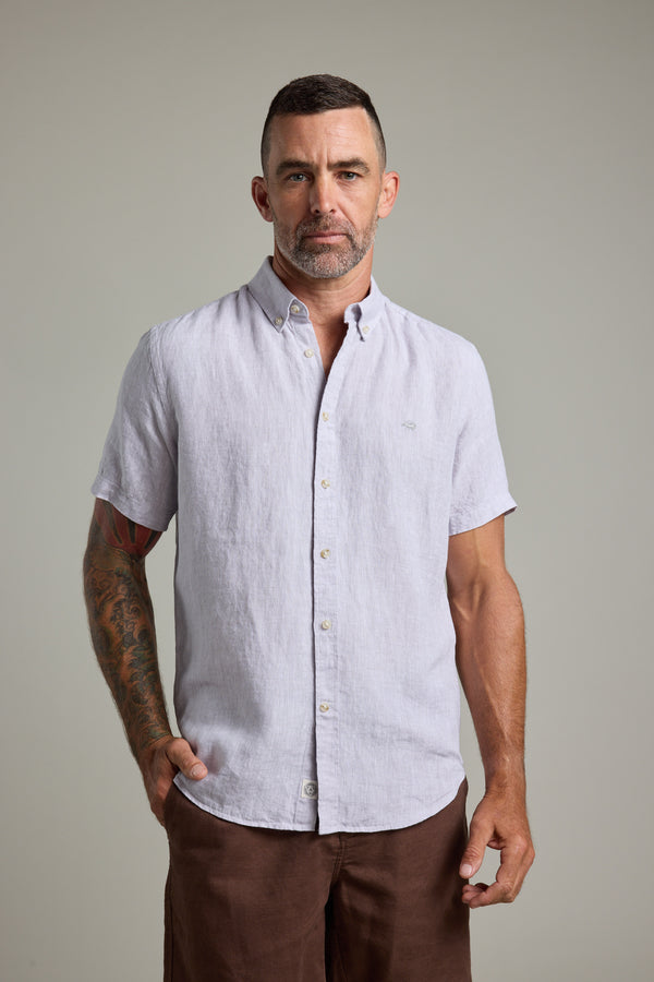 A man with short dark hair, a beard, and a tattoo sleeve on his left arm stands facing forward in Barkers' Gentry Linen Shirt (light gray, short-sleeved) and brown pants, wearing a neutral expression.