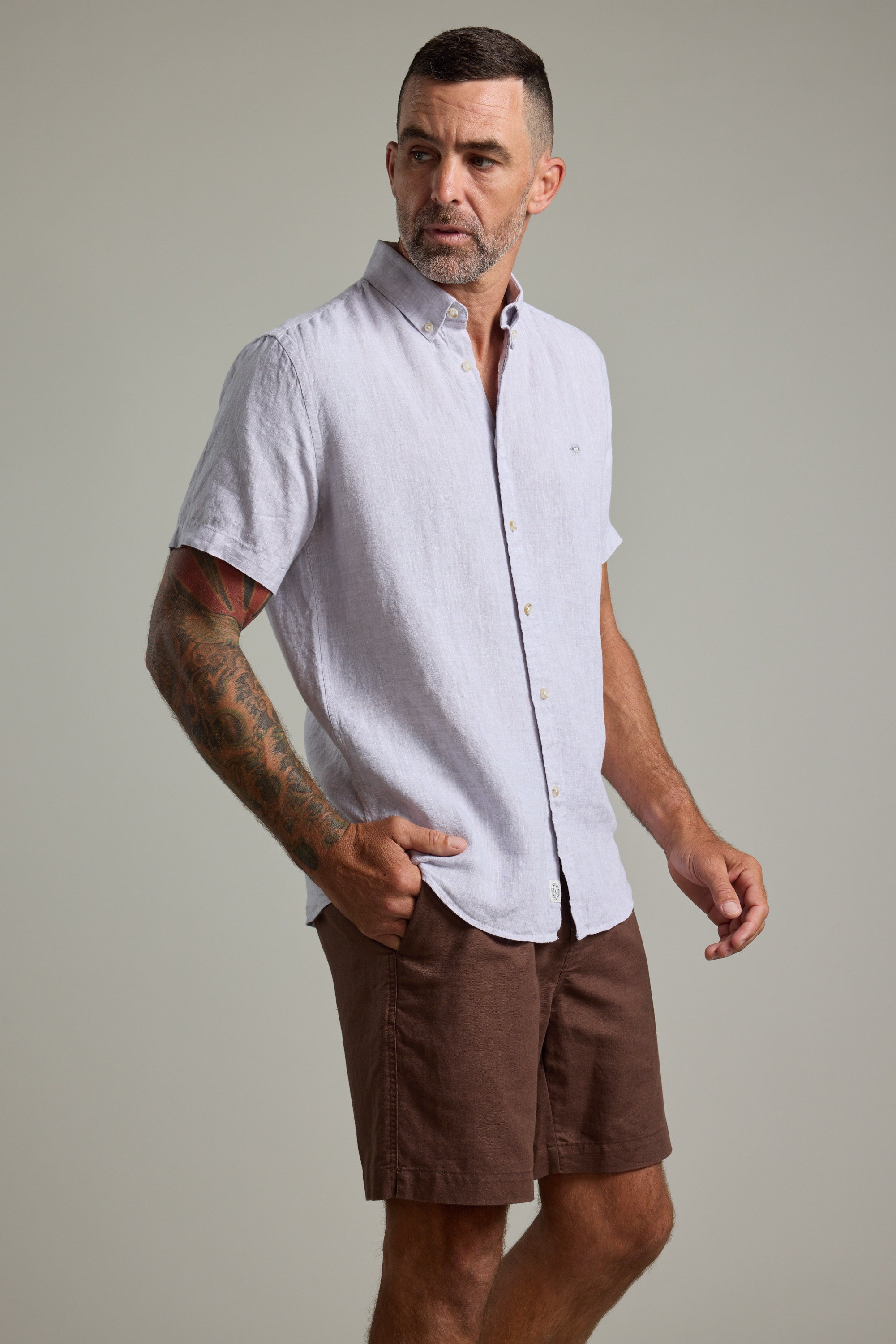 A man with short hair and a trimmed beard wears the Barkers Gentry Linen Shirt, a light short-sleeve button-up, paired with brown shorts. He stands with one hand in his pocket against a plain, neutral background, looking to the side.