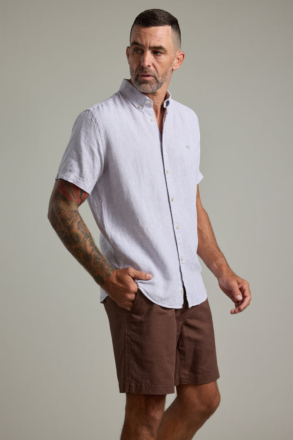 A man with short hair and a trimmed beard wears the Barkers Gentry Linen Shirt, a light short-sleeve button-up, paired with brown shorts. He stands with one hand in his pocket against a plain, neutral background, looking to the side.