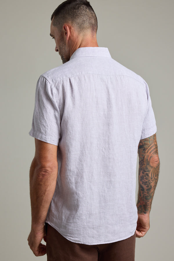 A man with short hair and a tattooed right arm stands with his back to the camera, wearing the Barkers Gentry Linen Shirt in a light shade and brown pants against a plain background.