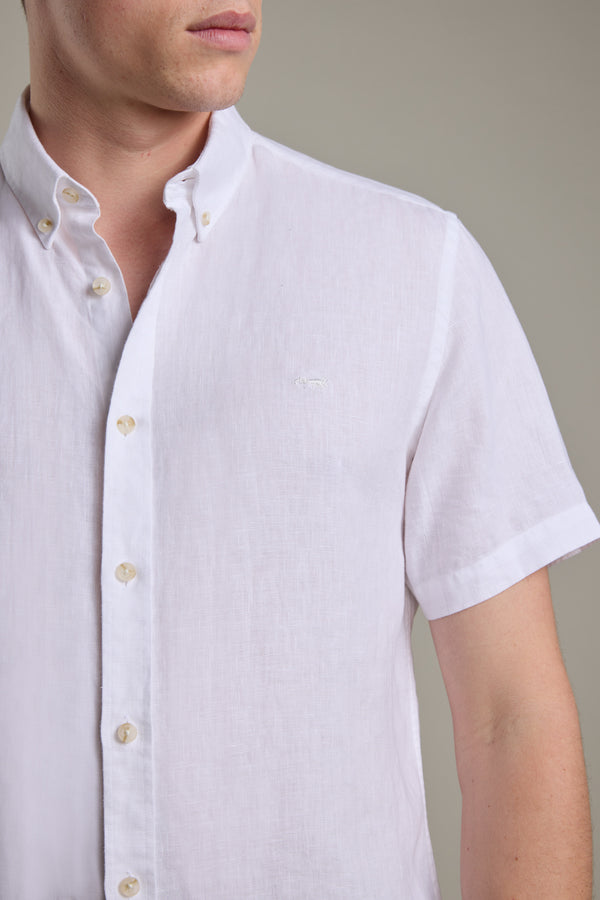 A person wears the Barkers Gentry Linen Shirt—a short-sleeved, white French linen button-up with a collar, light buttons, and a crayfish embroidery on the chest. The background is plain and neutral; their face is partially visible.