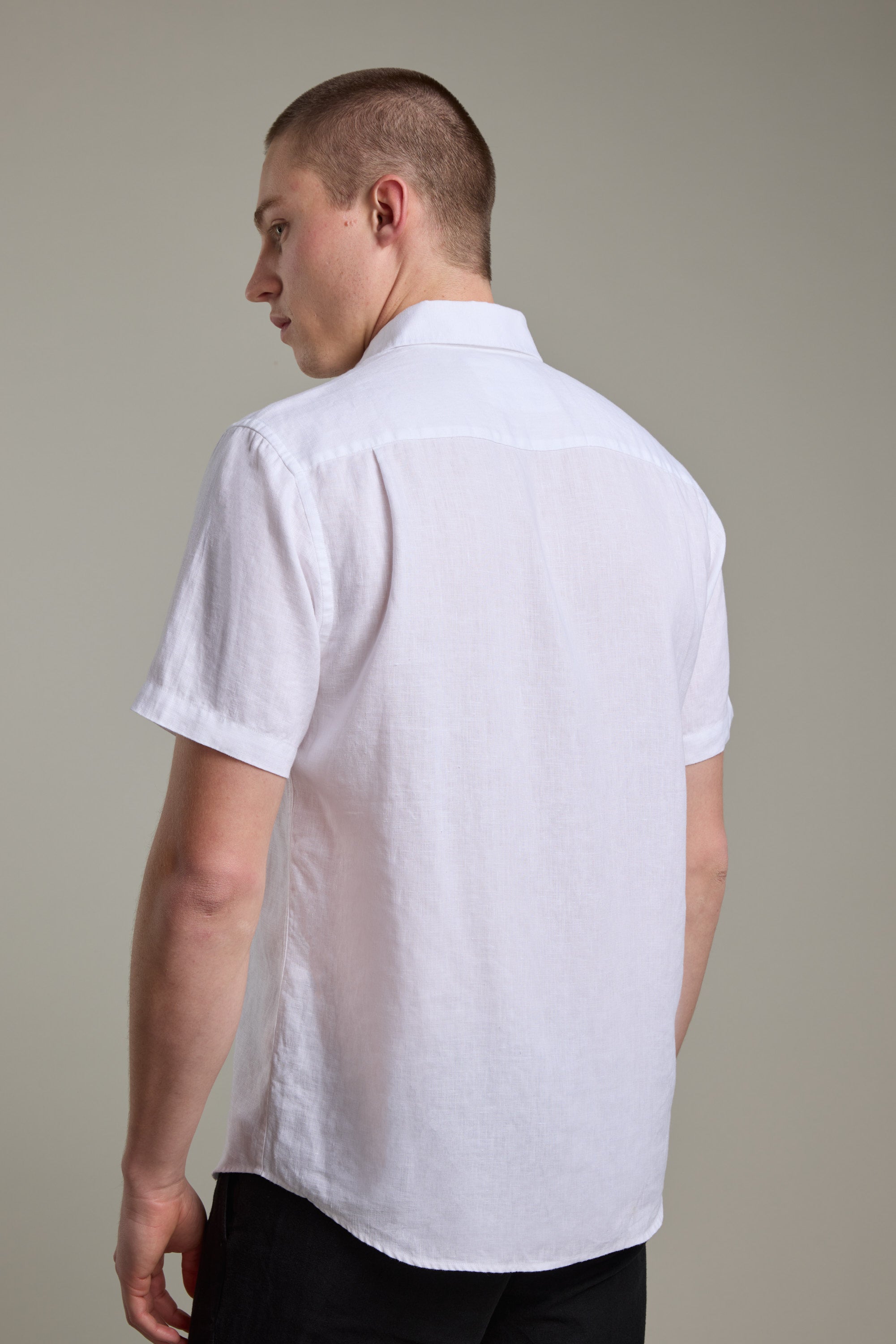 A person with short hair, seen from behind and looking over their left shoulder, wears the Barkers Gentry Linen Shirt—white, French linen, short-sleeve button-up—with black pants against a plain background.