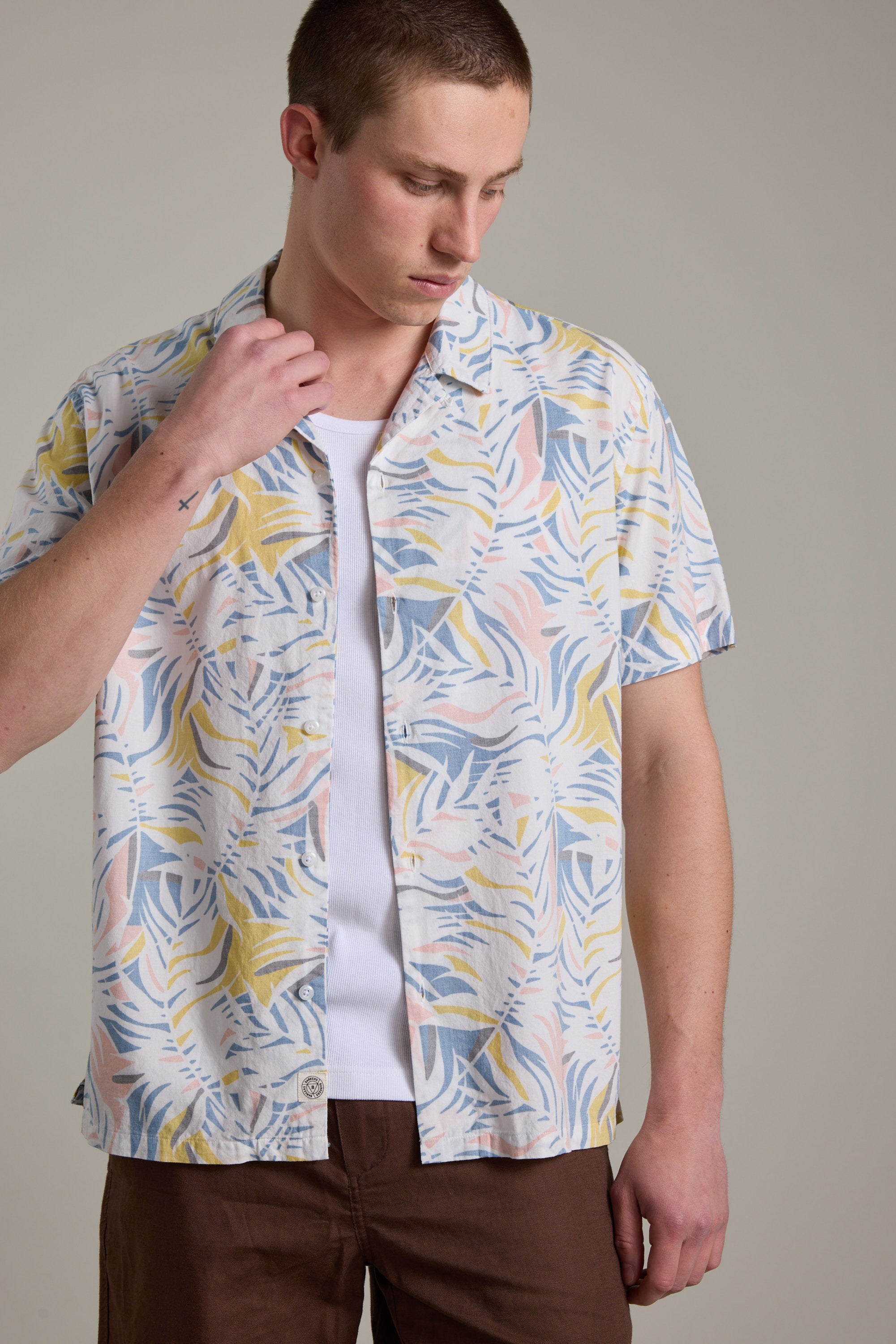 A person wears the Barkers Ribbed Singlet in white as a base, layered under a pastel tropical leaf short-sleeve shirt and brown pants, standing against a plain light background and looking down.