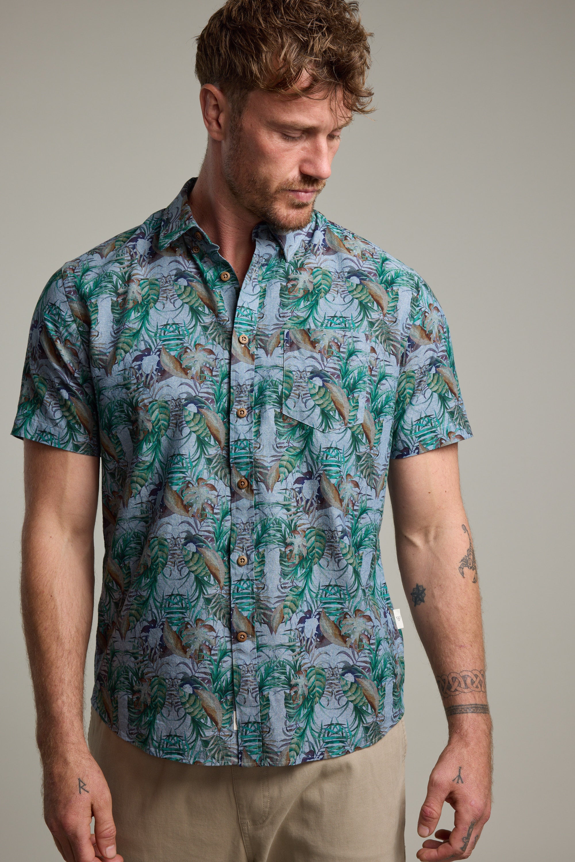 A man with short, curly brown hair and a tattooed left arm models the Barkers Rockport Print Shirt—a green and blue tropical leaf cotton shirt—against a plain background, glancing down to his left.