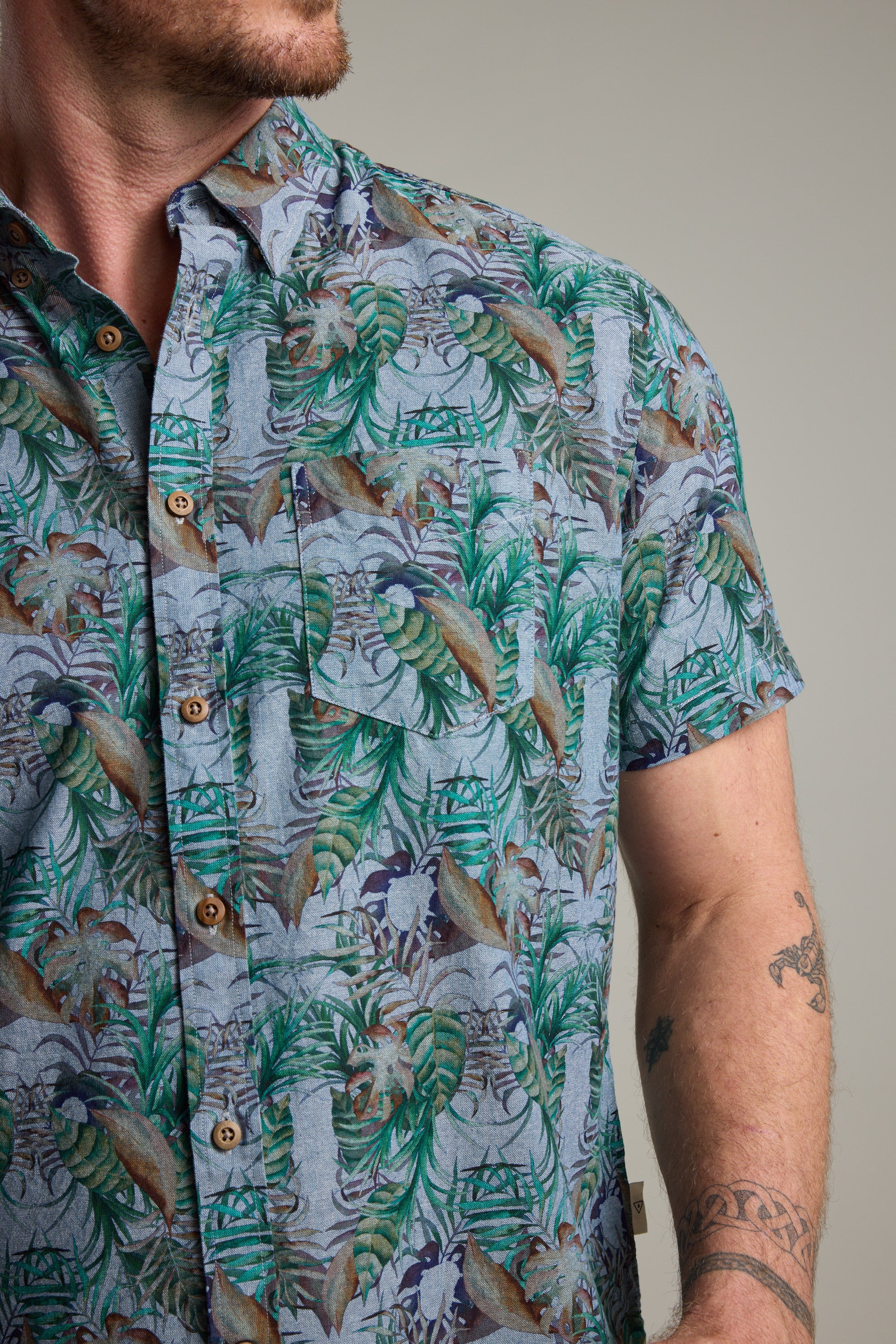 A man wears a Barkers Rockport Print Shirt with a vibrant tropical leaf design, ideal for summer parties. The focus is on the bold print and his tattooed arm, with his face partially out of frame.