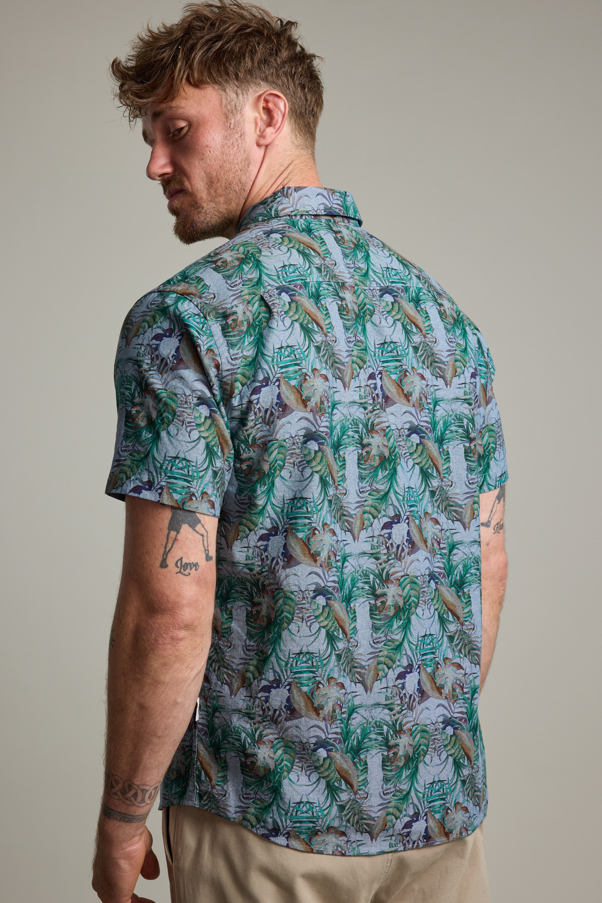 A man with short, wavy hair and tattooed arms models the Barkers Rockport Print Shirt, a vibrant tropical summer party shirt. He stands slightly turned to showcase the colorful back against a plain background.