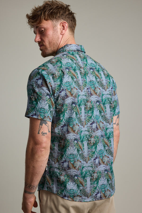 A man with short, wavy hair and tattooed arms models the Barkers Rockport Print Shirt, a vibrant tropical summer party shirt. He stands slightly turned to showcase the colorful back against a plain background.