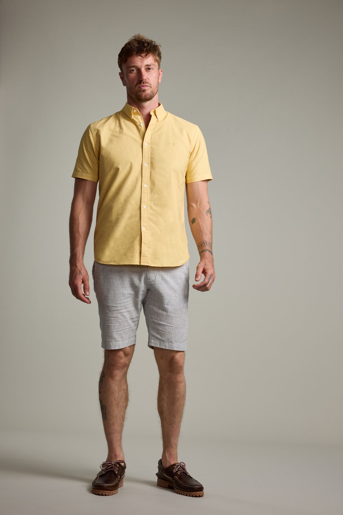 A man faces forward wearing a yellow Parker Oxford Shirt by Barkers, made from responsibly sourced cotton, paired with light gray shorts and brown shoes. The background is plain and neutral.