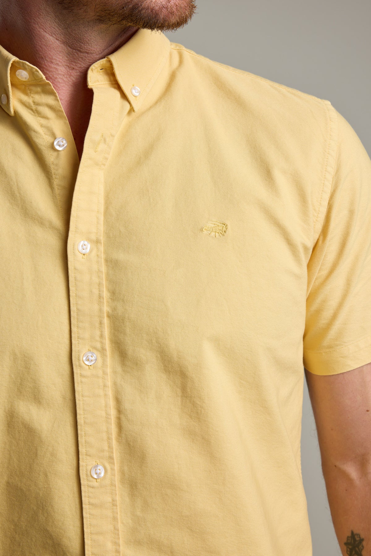 A man wears a Barkers Parker Oxford Shirt in light yellow with a classic fit and small embroidered logo. Made from responsibly sourced cotton, the photo shows his torso and part of his face against a neutral background.