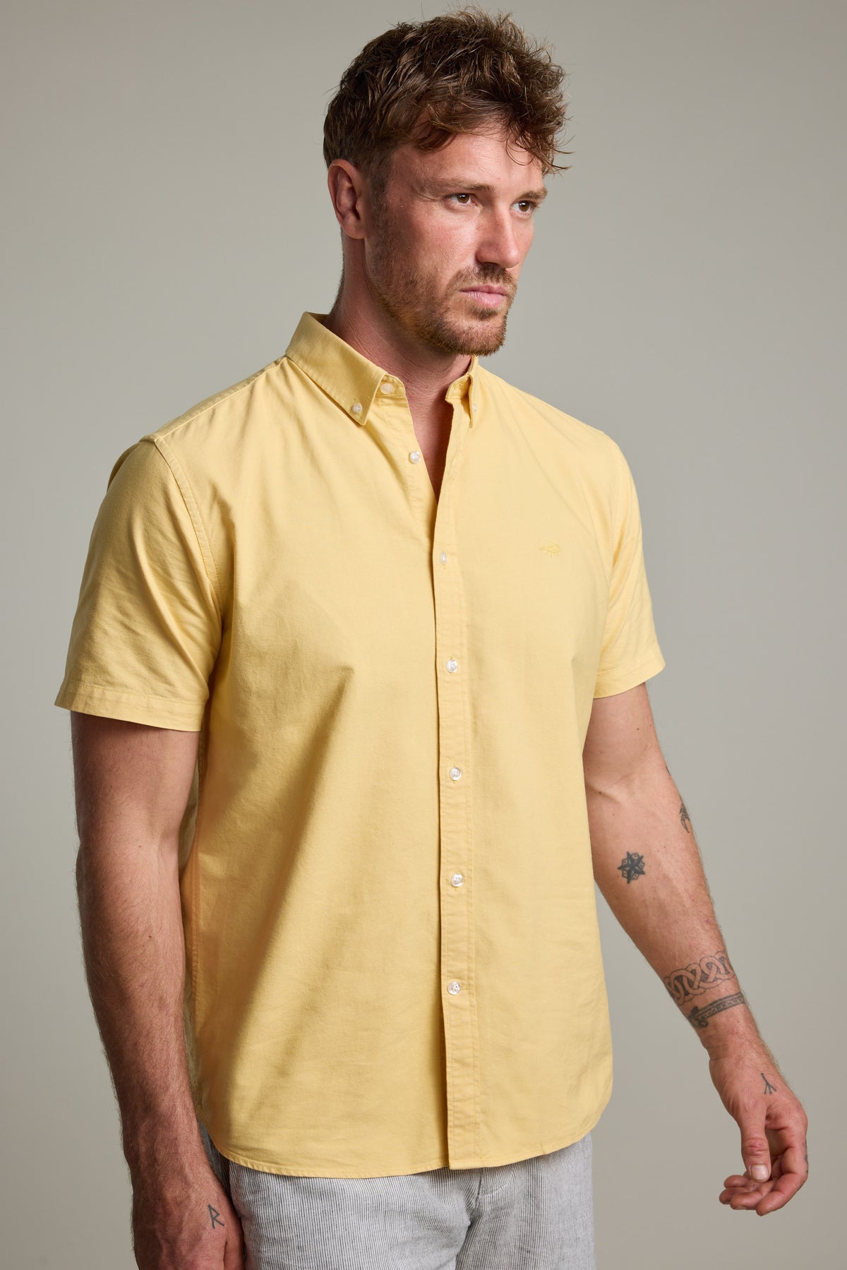 A man with short brown hair and facial hair wears a light yellow Parker Oxford Shirt in classic fit by Barkers, made from responsibly sourced cotton, with light gray pants. He has tattoos on his arms and stands against a plain, light background.