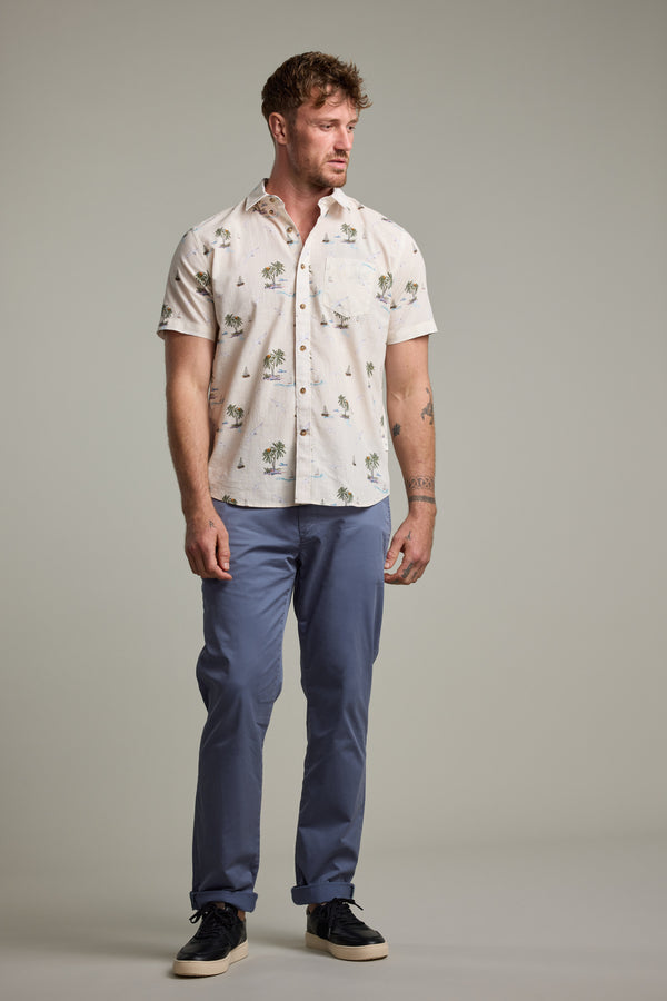 A man stands against a plain background wearing the Barkers Island Print Shirt, blue pants, and black sneakers, looking slightly to the side with a relaxed posture that captures effortless summer style.