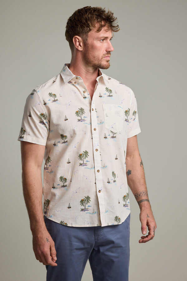 A man models the Barkers Island Print Shirt, a short sleeve, light-colored shirt featuring palm trees and sailboats. He pairs it with blue pants for a relaxed summer look.