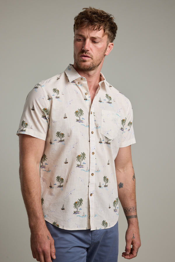 A man with tousled hair models the Barkers Island Print Shirt, a light-colored short sleeve shirt with palm trees and sailboats, standing against a plain background and gazing left for an effortlessly relaxed summer look.