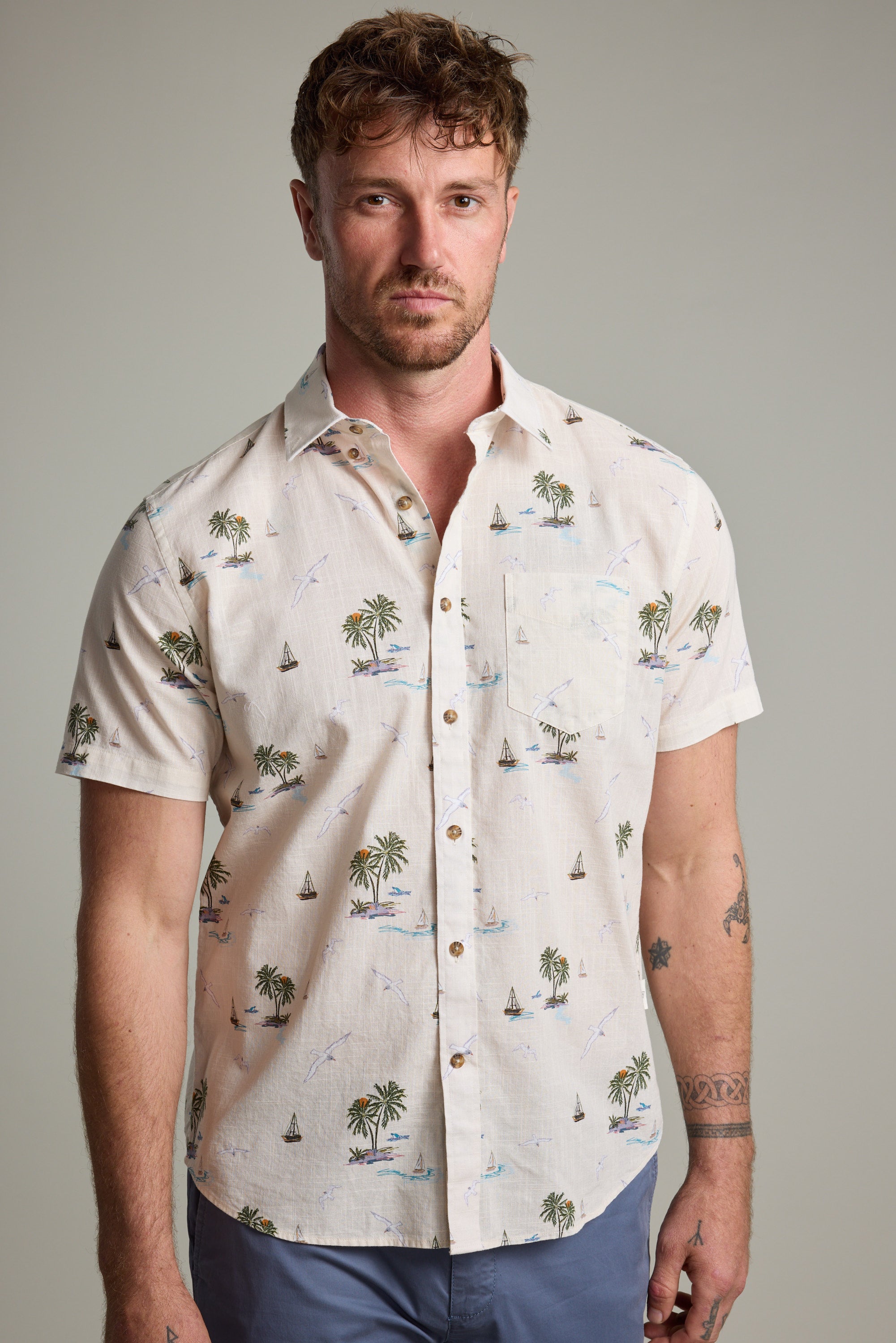 A man with short curly hair and a light beard models the Barkers Island Print Shirt—short-sleeve, light-colored, featuring palm trees and sailboats—styled with blue pants against a plain background.