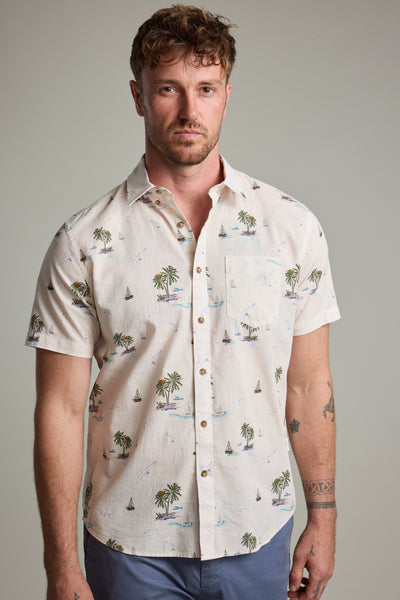 A man with short curly hair and a light beard models the Barkers Island Print Shirt—short-sleeve, light-colored, featuring palm trees and sailboats—styled with blue pants against a plain background.