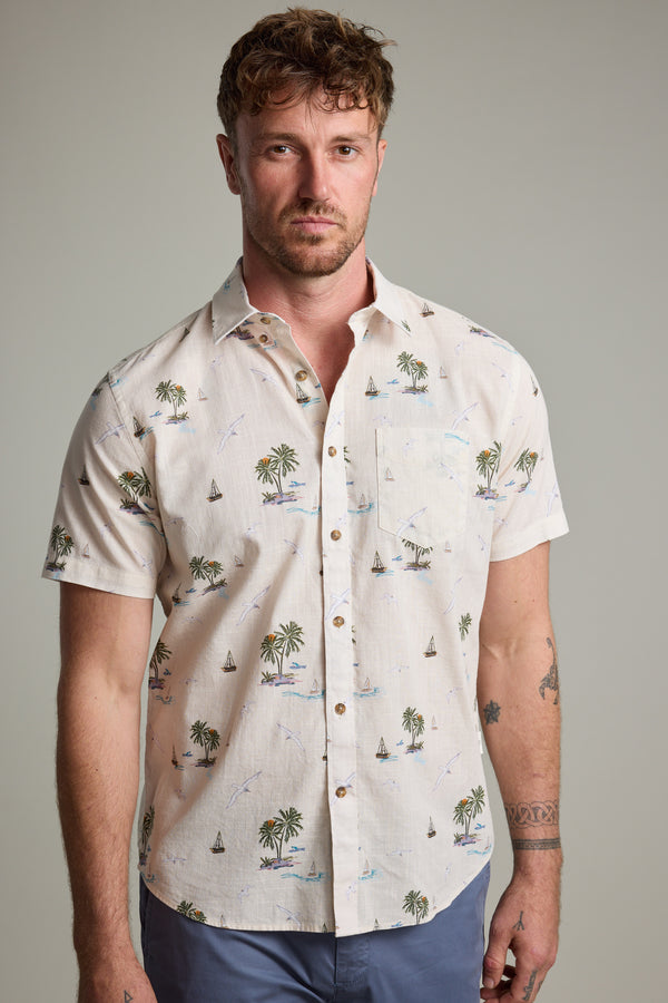 A man with short curly hair and a light beard models the Barkers Island Print Shirt—short-sleeve, light-colored, featuring palm trees and sailboats—styled with blue pants against a plain background.