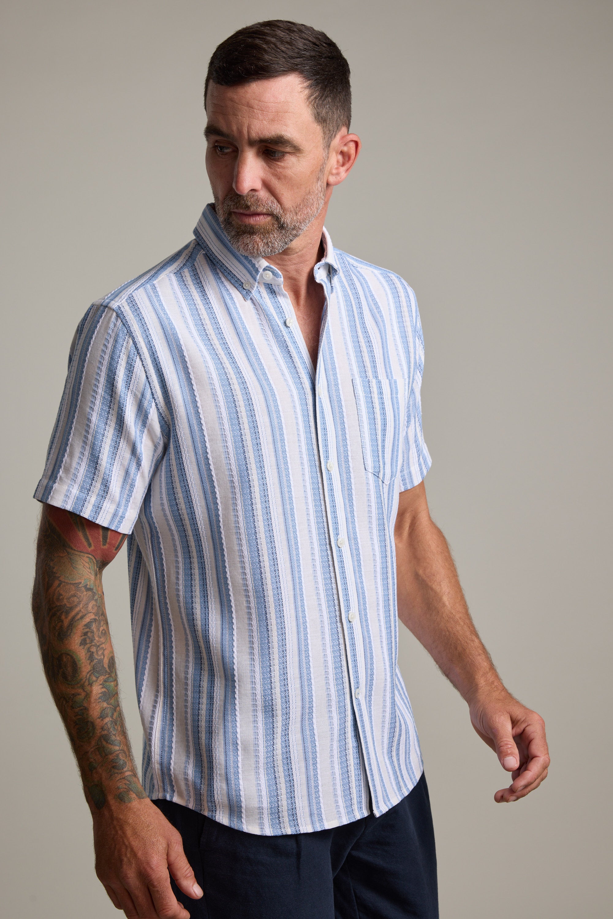 A man with short dark hair and a beard wears the Barkers Merten Stripe Shirt—a short-sleeve, light blue and white striped summer staple. He has a tattoo on his left arm and stands against a plain background, looking to his left.