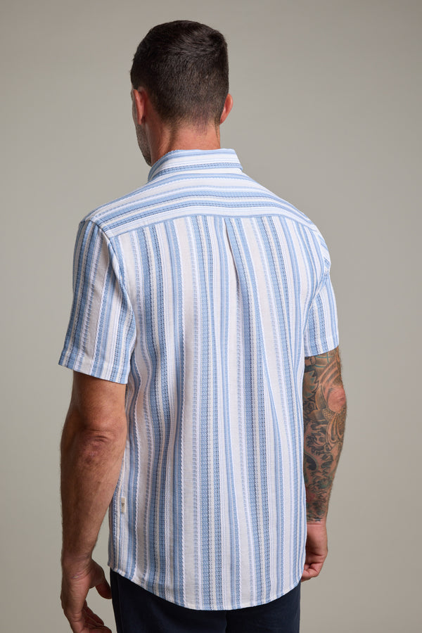 A man with short dark hair and a tattooed right arm is seen from the back wearing the Barkers Merten Stripe Shirt, featuring vertical blue and white stripes—an ideal cotton piece for any summer wardrobe, set against a plain background.