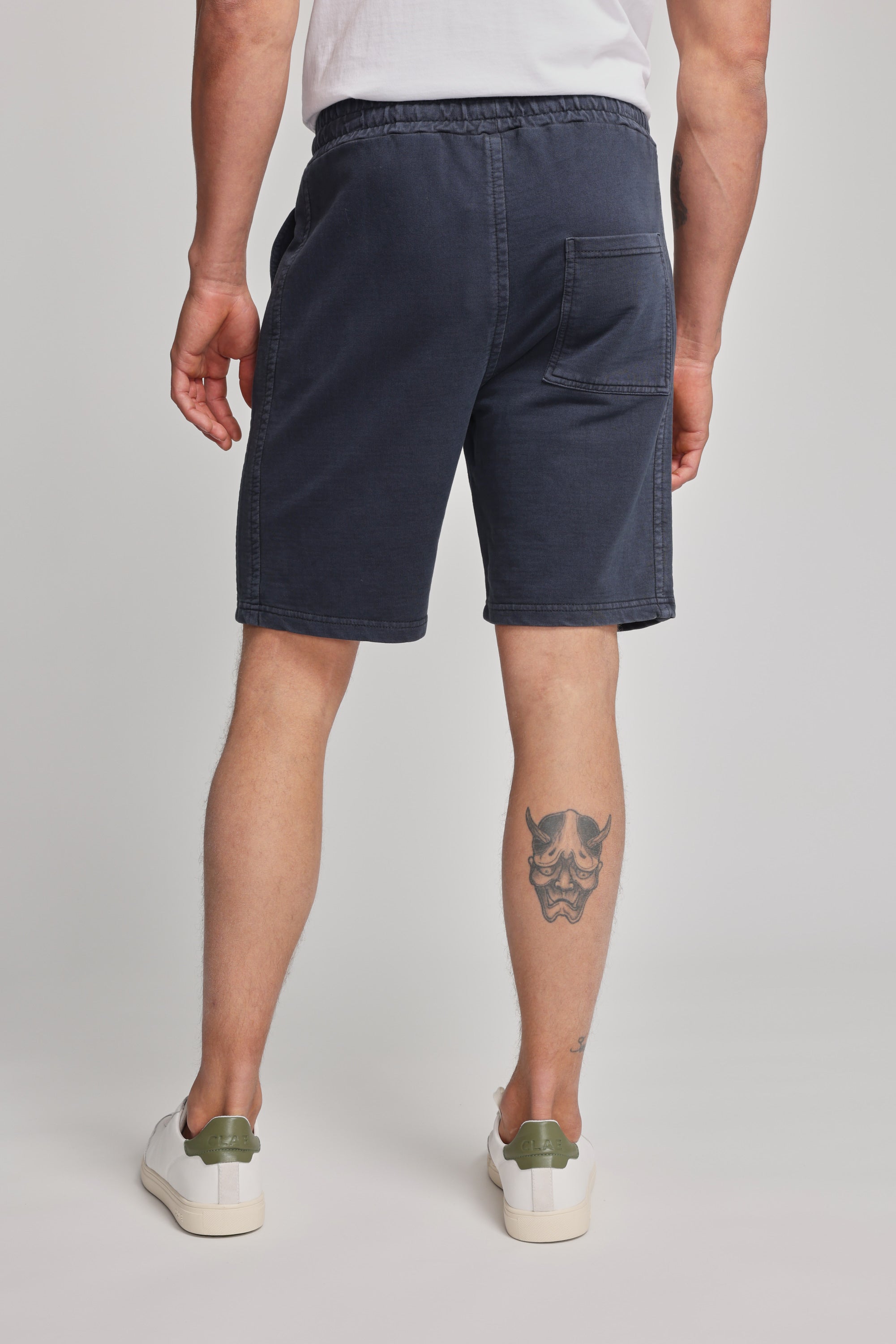 A person shown from the back wears a white shirt, Barkers Organic Classic Print Track Short in navy blue, and white sneakers with green accents. A stylized face tattoo appears on their left calf. The shorts display the Barkers heritage logo.