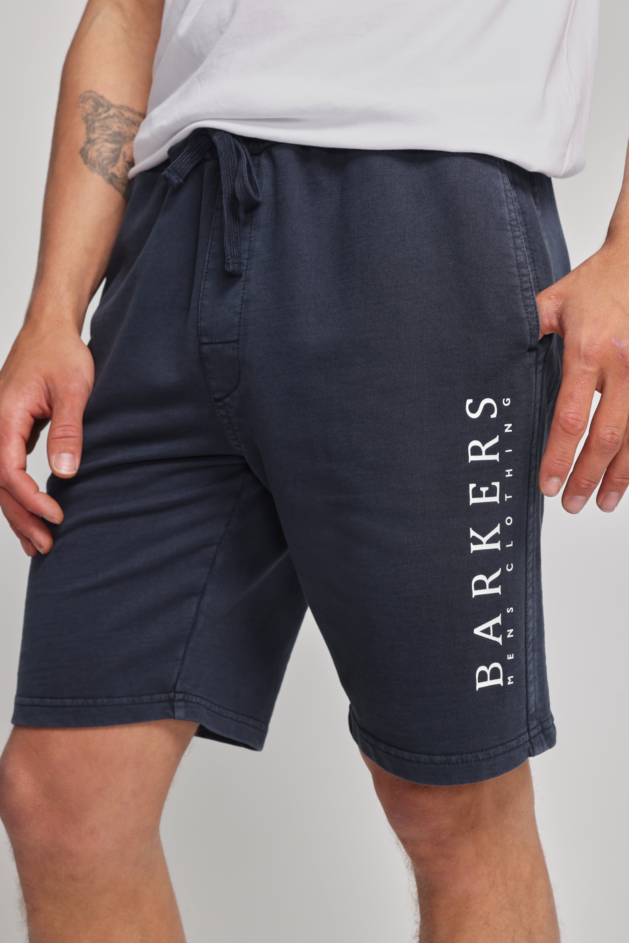 A person wears Barkers Organic Classic Print Track Short in navy, featuring a vertical heritage logo on the left leg, paired with a white t-shirt. Only the lower torso and upper legs are shown.