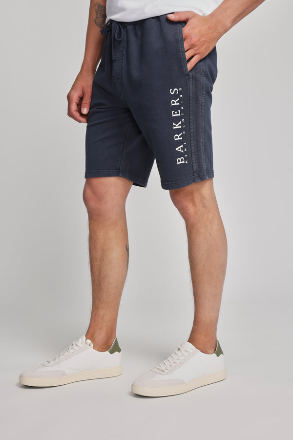 Wearing Barkers Organic Classic Print Track Shorts with the heritage logo on the left leg, a white t-shirt, and white sneakers with green accents, a person stands against a plain light background.