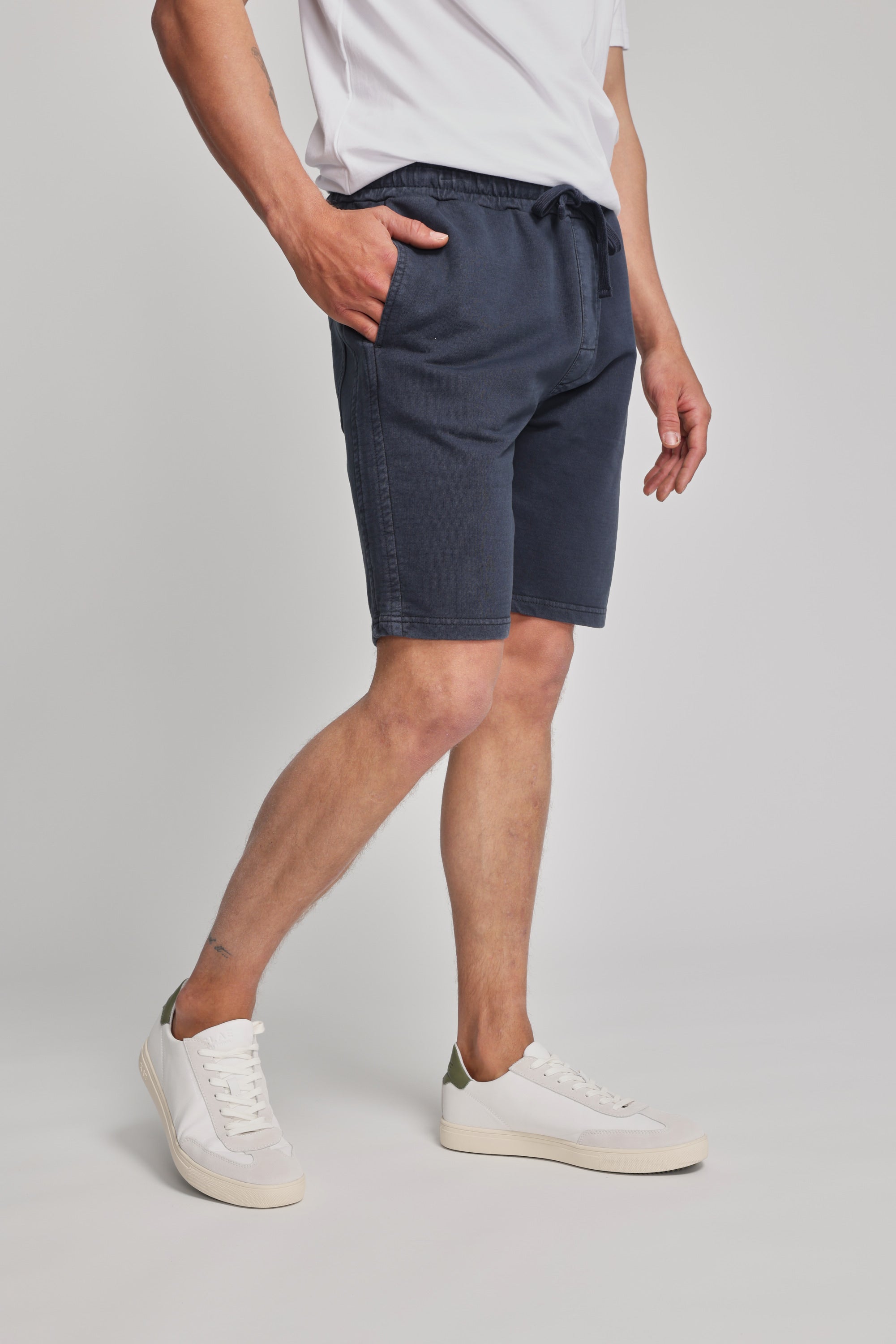 Someone wearing a white t-shirt, Barkers Organic Classic Print Track Short, and white sneakers stands with one hand in their pocket against a plain light background.