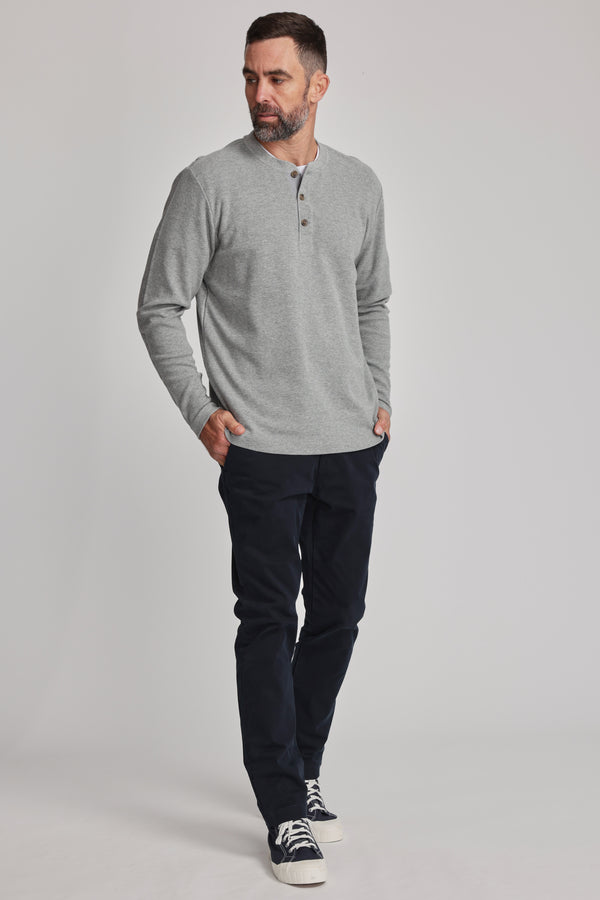 A man with a beard stands against a plain background, wearing the Barkers Avenue Waffle Henley in light gray, paired with dark navy pants and black-and-white sneakers, hands in his pockets.