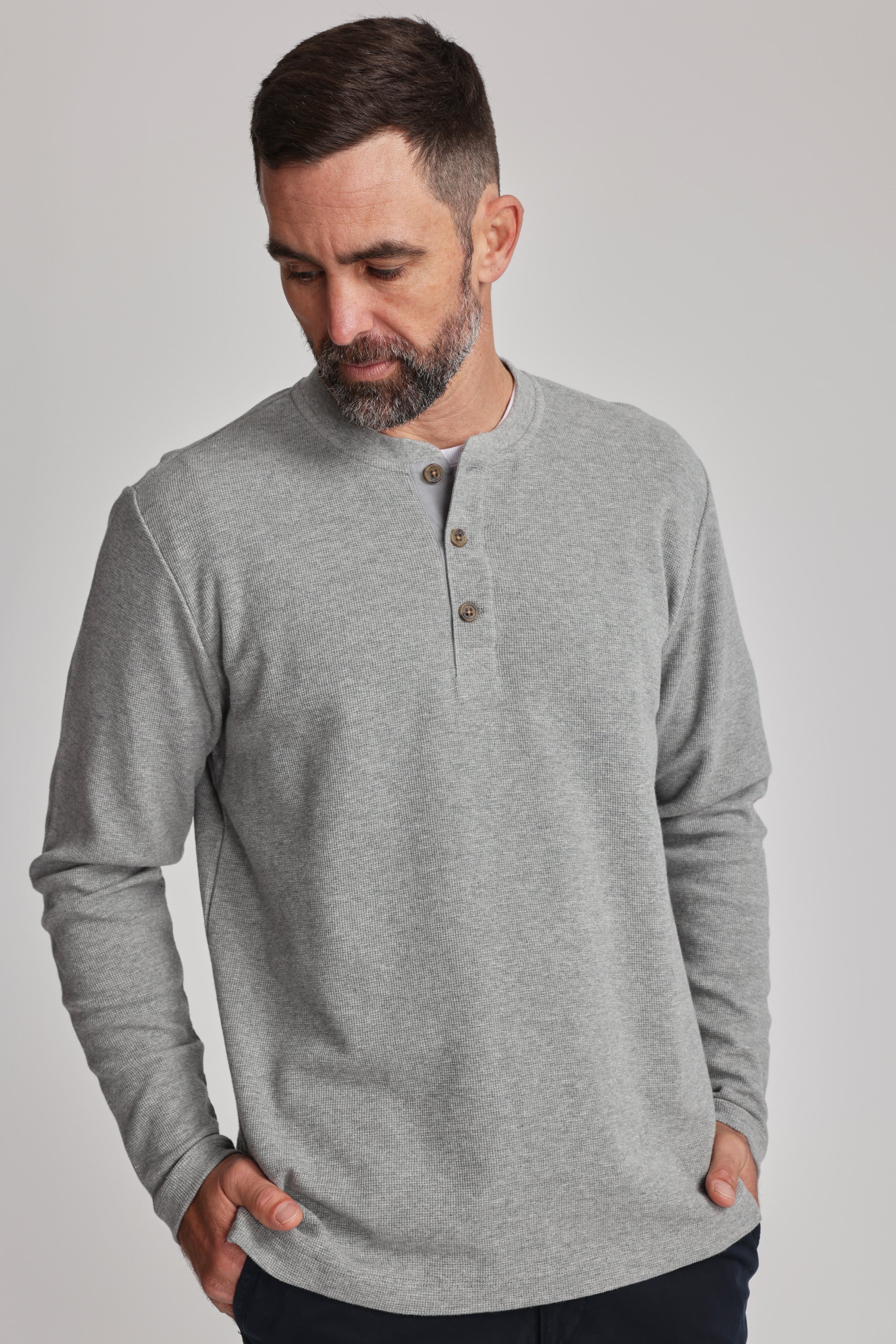 A man with short dark hair and a beard wears the Barkers Avenue Waffle Henley, a mid-weight organic cotton shirt with a subtle waffle texture. He looks down, hands in his pockets, against a plain light background.