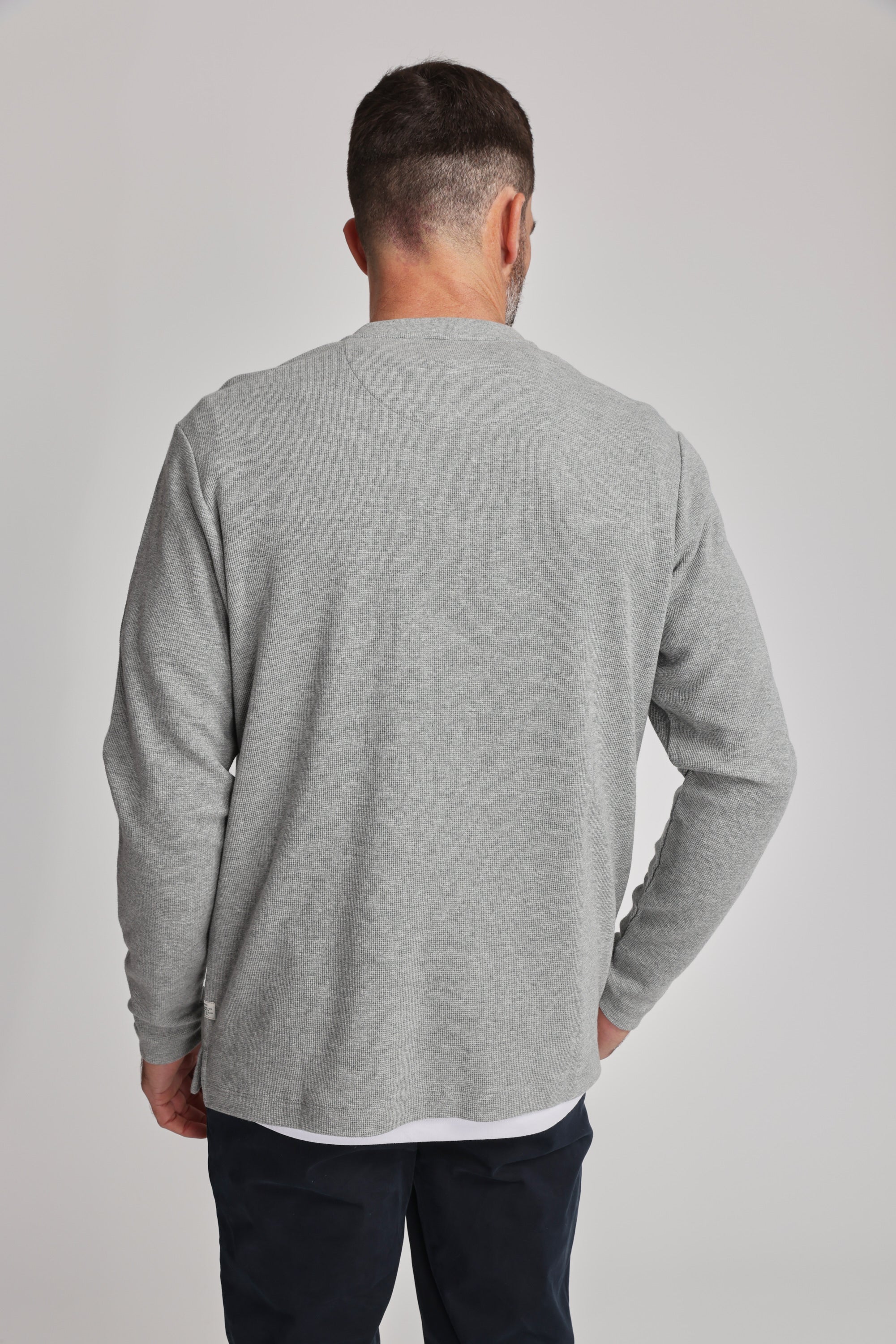 A man with short dark hair and a trimmed beard stands with his back to the camera, wearing the Barkers Avenue Waffle Henley in light gray and dark pants against a plain light background.