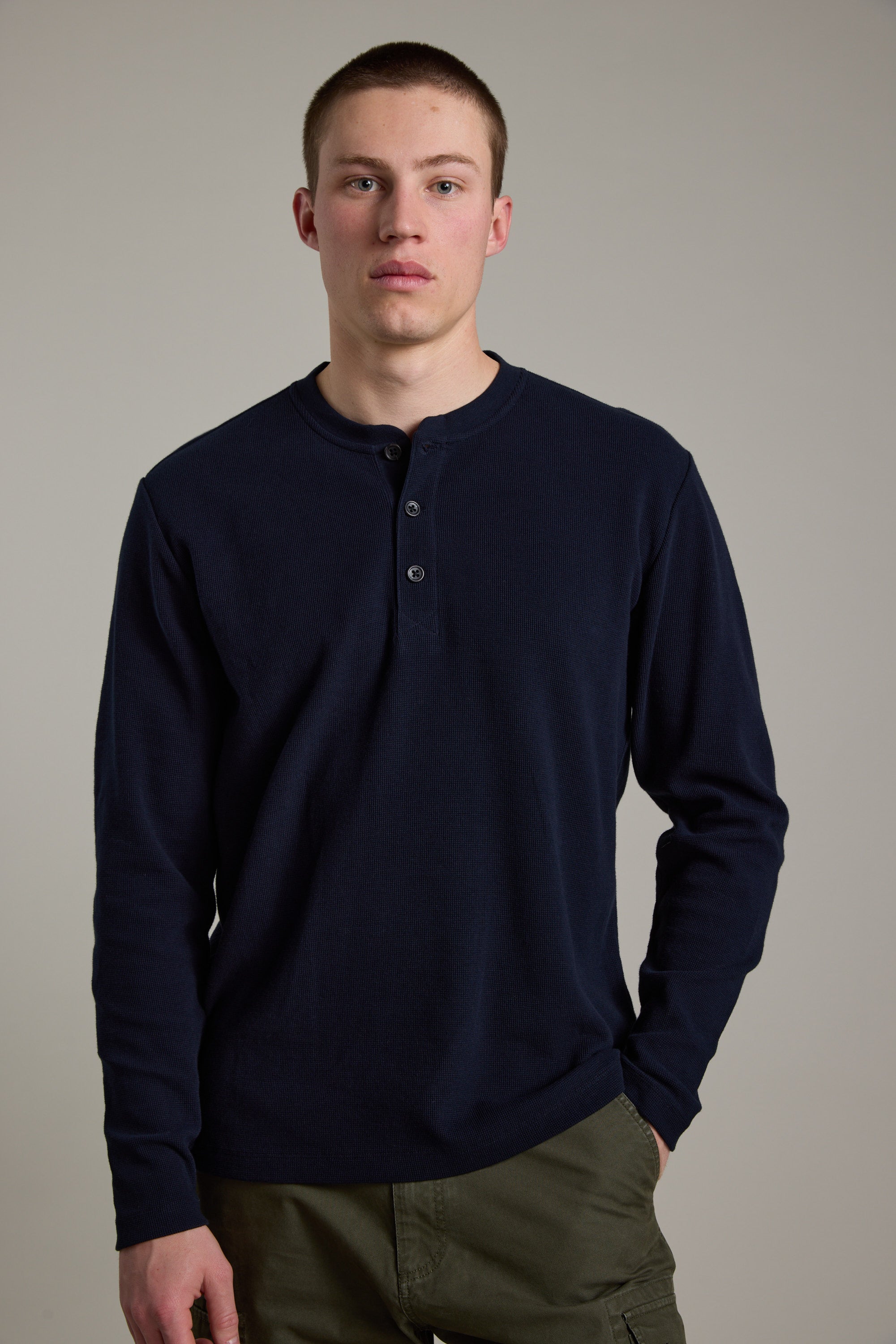 A young man with short hair wears the Barkers Avenue Waffle Henley in navy blue, made from organic cotton, and olive green pants, standing against a plain light gray background.