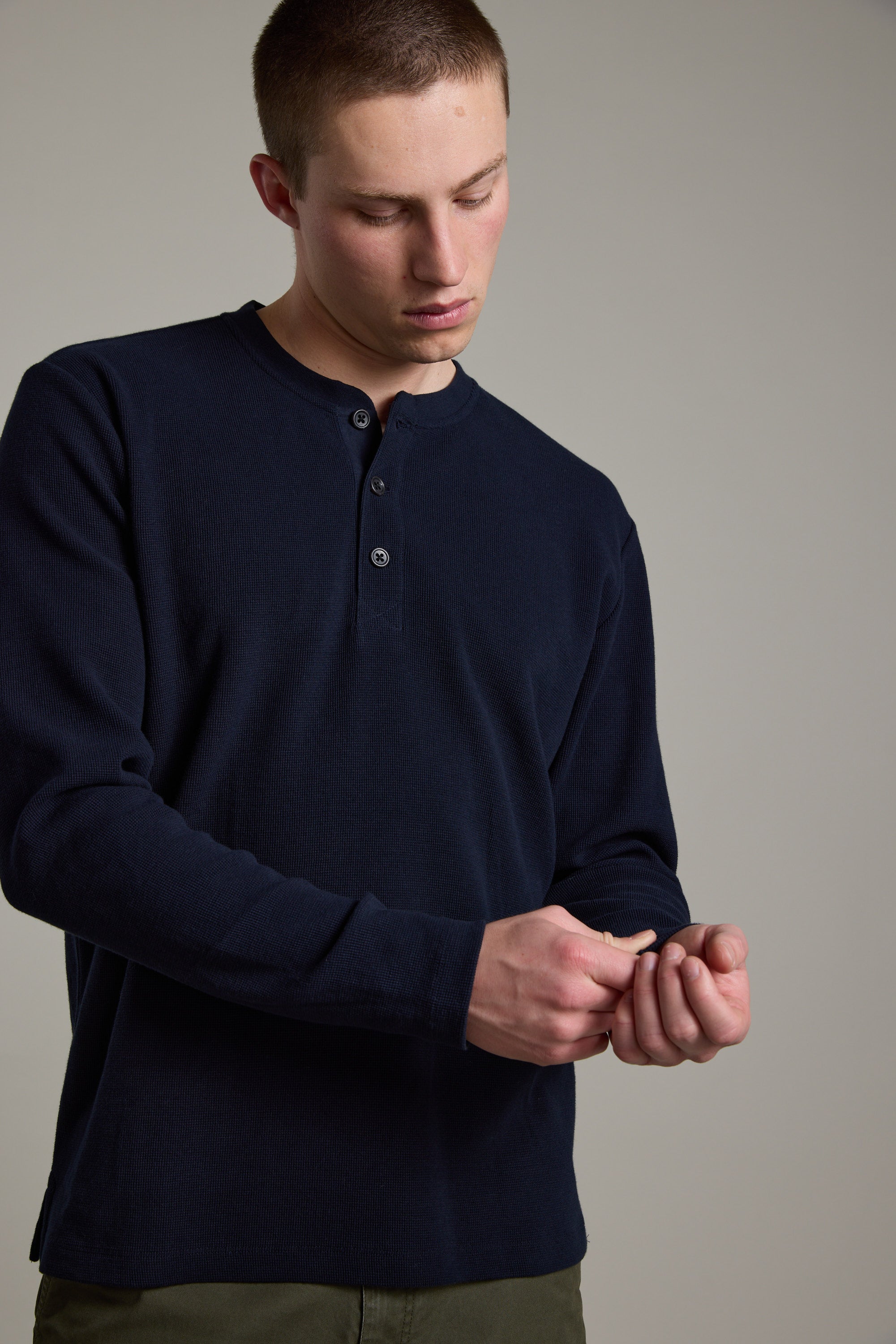 A young man wears the Barkers Avenue Waffle Henley in dark navy—an organic cotton, mid-weight top—and olive green pants, adjusting his cuff against a plain, light gray background.