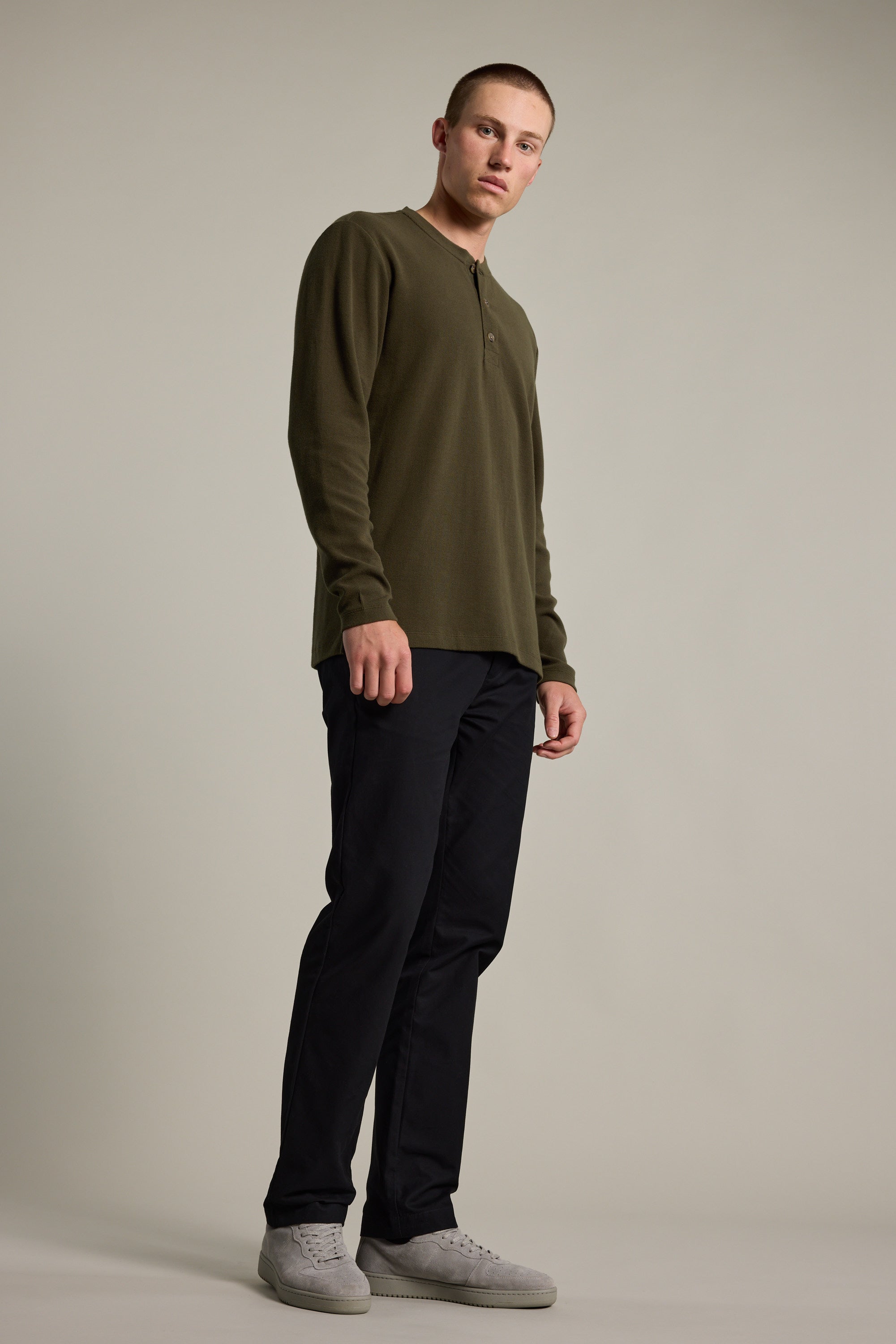 A young man stands against a plain backdrop in the Barkers Avenue Waffle Henley—an olive green organic cotton long-sleeve top—paired with dark pants and light gray sneakers. Ideal as a mid-weight layer for any season, he looks off to the side with a neutral expression.