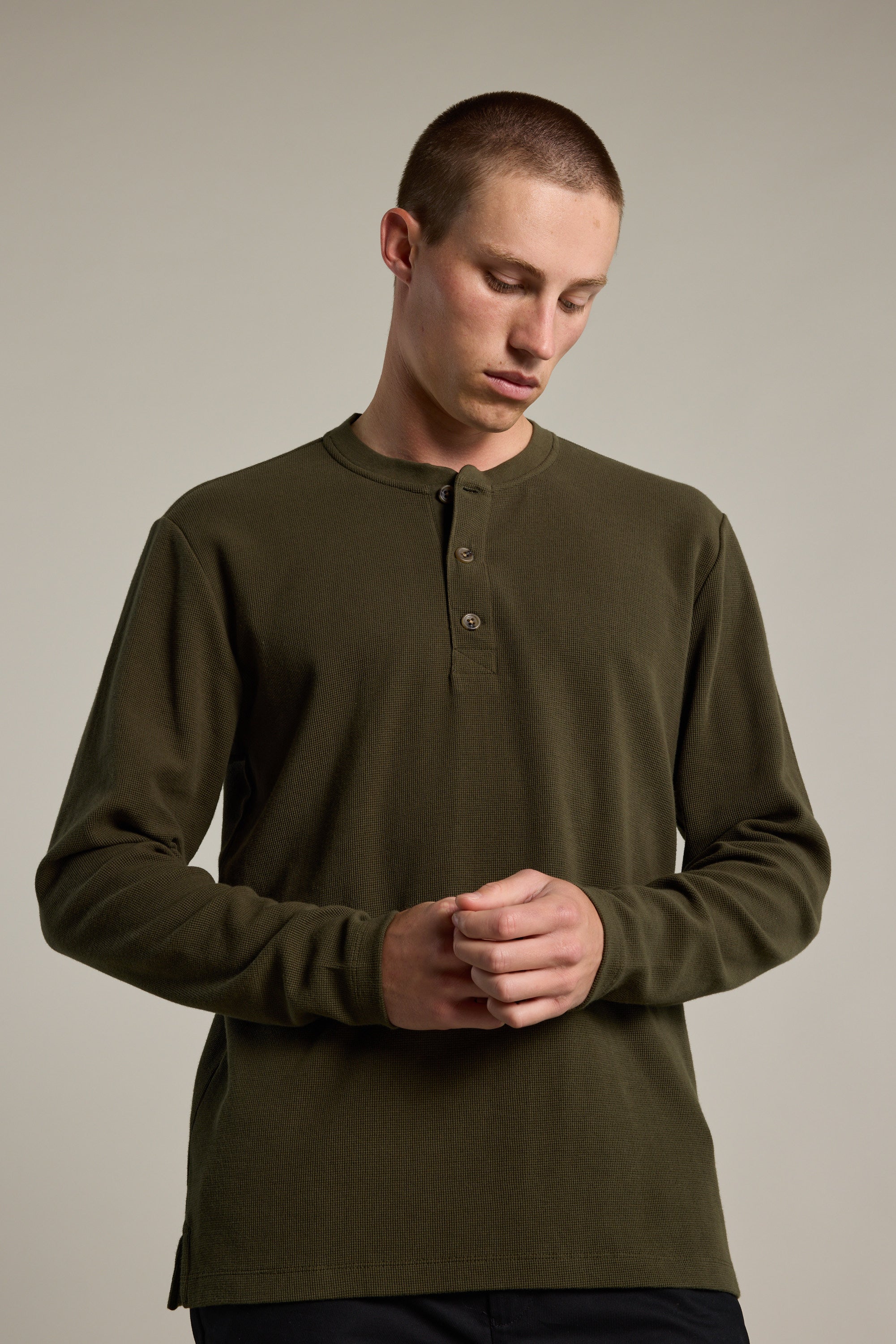 A young man with short hair models the Barkers Avenue Waffle Henley in olive green, made from organic cotton, standing against a light gray background and looking down with his hands loosely clasped in front of him.